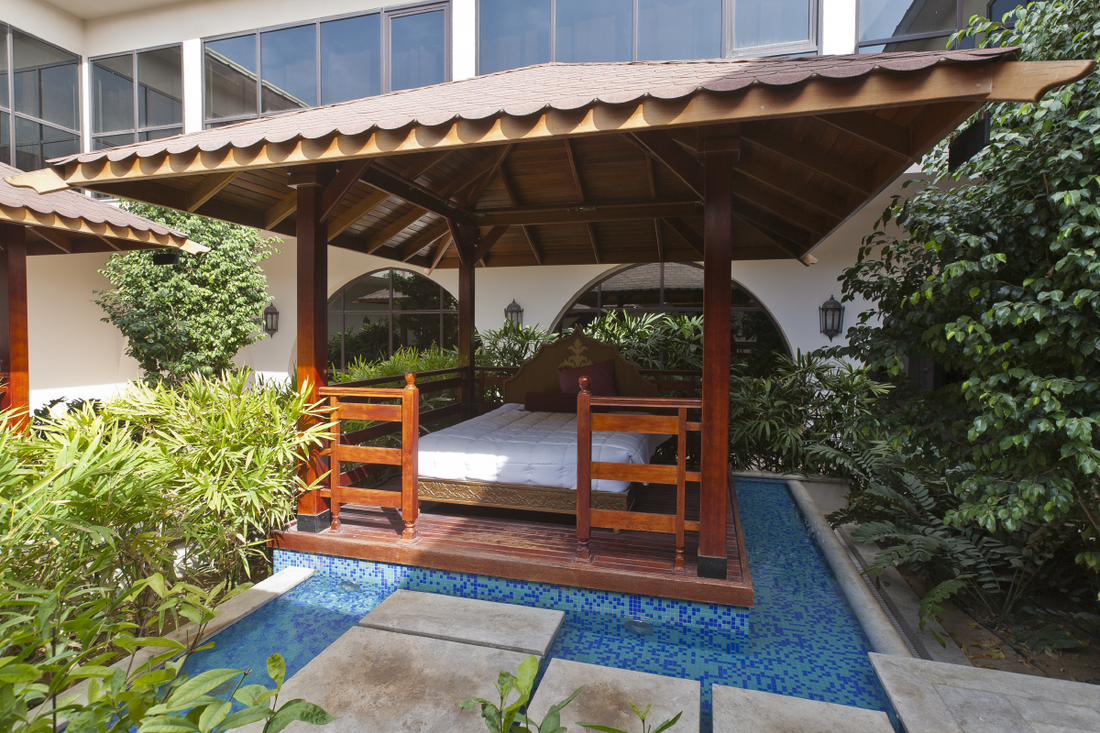 Wooden pergola with bed and railing over blue tiled water feature surrounded by greenery in hotel courtyard