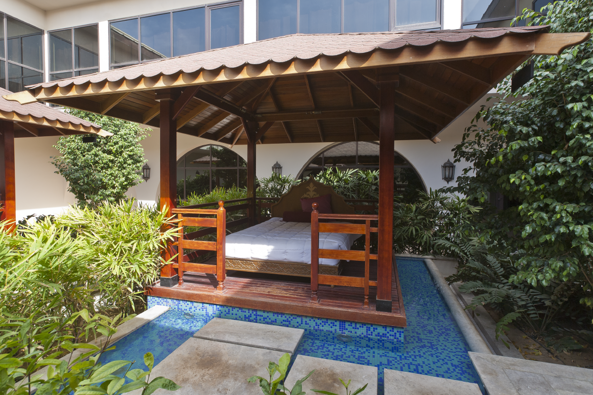 Wooden pergola with bed and railing over blue tiled water feature surrounded by greenery in hotel courtyard