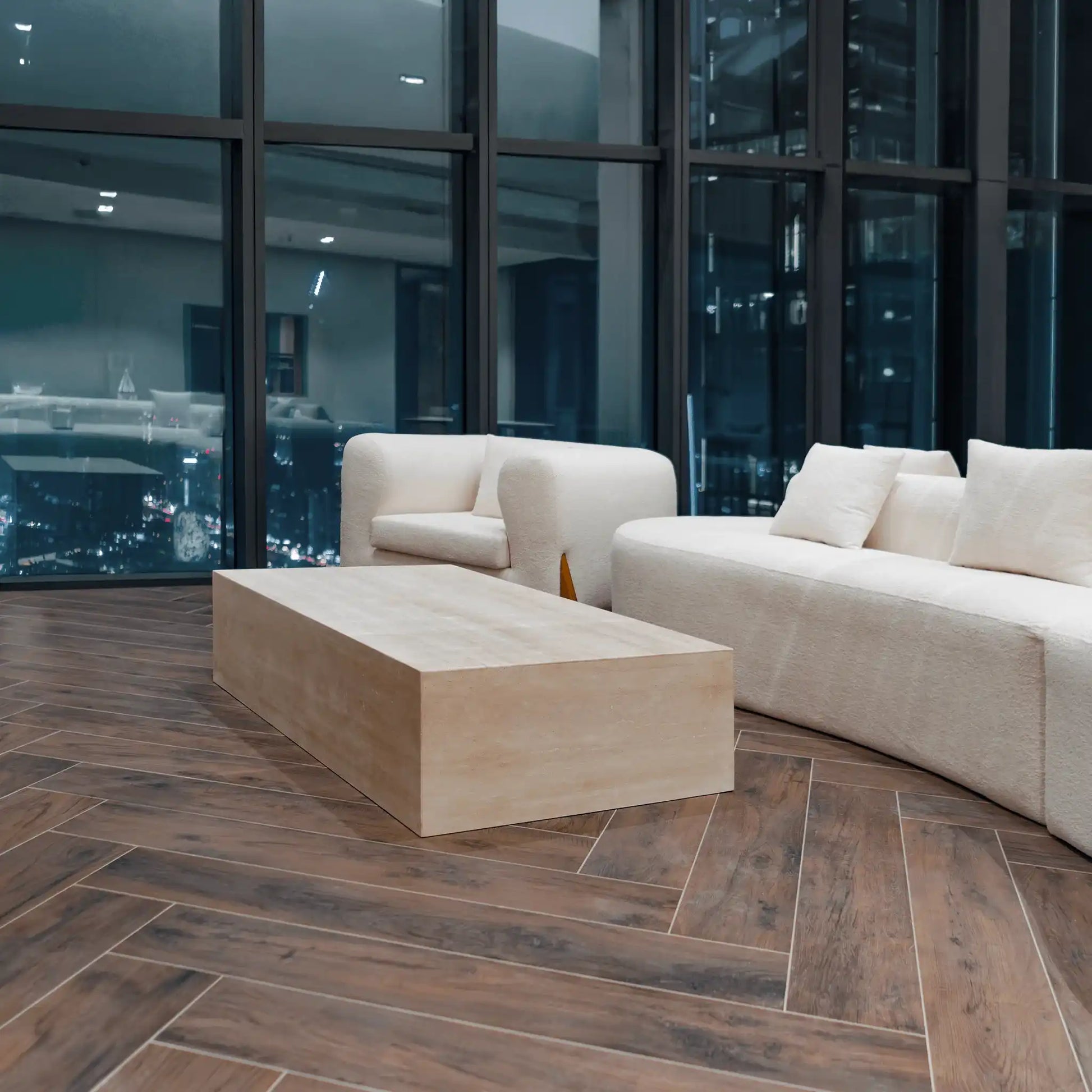 Modern living room with a beige sofa and Linea travertine coffee table from Marbeya Charm in front of large windows with cityscape view.