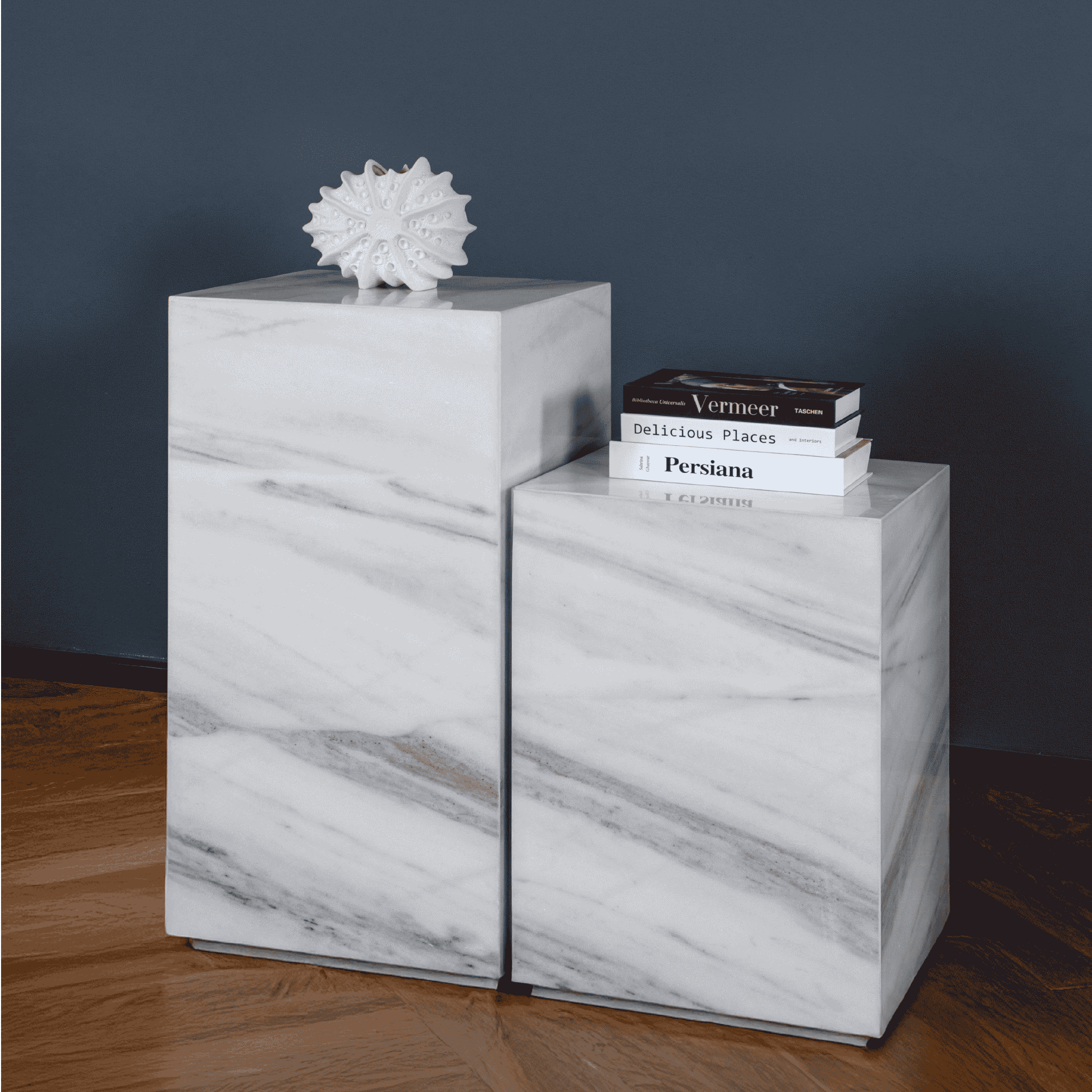 Two white marble  plinths with a decorative object and books on a wooden floor.